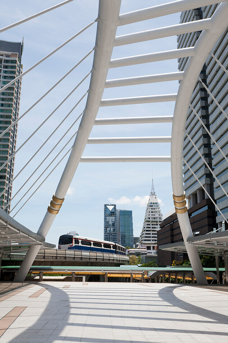 A mass transit train on an elevated track passes modern buildings in the central business district of Bangkok. Public transport. A passenger bridge and covered walkway., Bangkok, Thailand/ Above Ground train