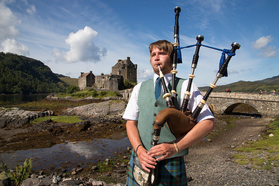 Junger Mann im Schottenrock spielt Dudelsack vor Burg Eilean Donan Castle am Ufer von Loch Duich, nahe Dornie, Highland, Schottland, Großbritannien, Europa