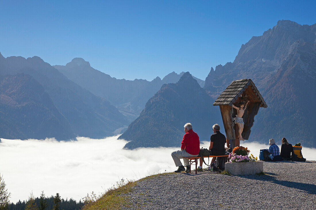 Hikers resting at a wayside cross, view over the fog in the valley onto Hochkalter, Berchtesgaden region, Berchtesgaden National Park, Upper Bavaria, Germany, Europe