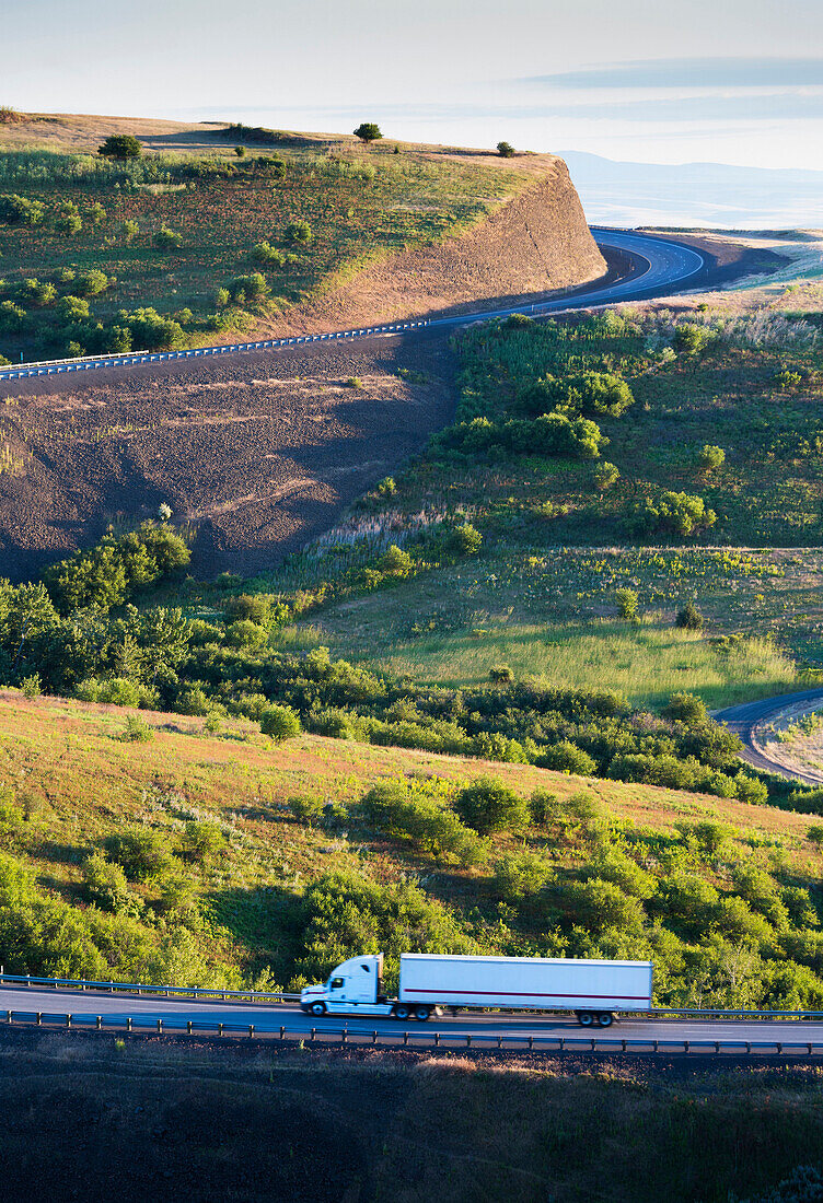 The landscape around the Interstate I-84 near Pendleton, Oregon, USA. A sharp bend in the road. Trees and grassland. An escarpment. Viewed from above. A truck travelling along the road.