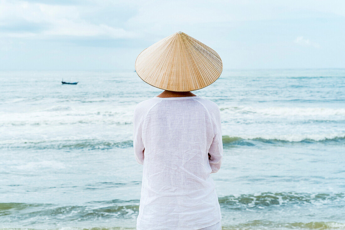 Frau mit vietnamesischer Kleidung und Strohhut blickt auf das Meer mit Fischerbooten, Strand von Mui Ne, Südvietnam, Vietnam, Asien
