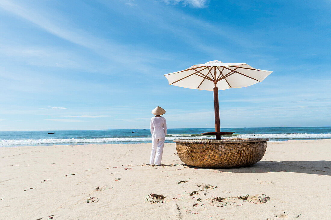 Woman wearing typical clothes and straw hat looking out to sea towards fishing boats, coast of Mui Ne, south Vietnam, Vietnam, Asia