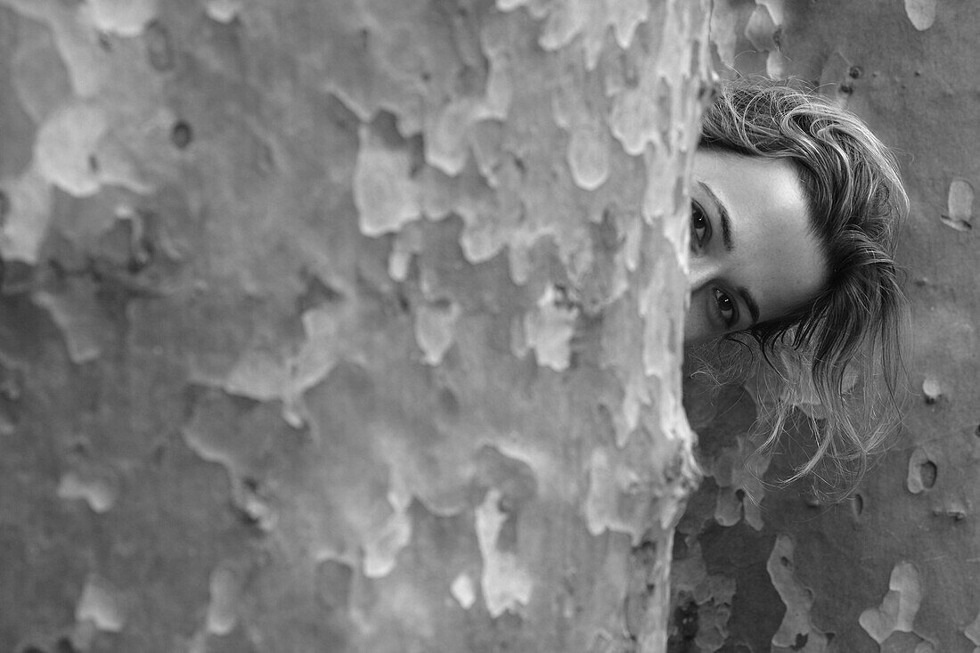 Young Woman Peeking Out From Behind Large Tree Trunk