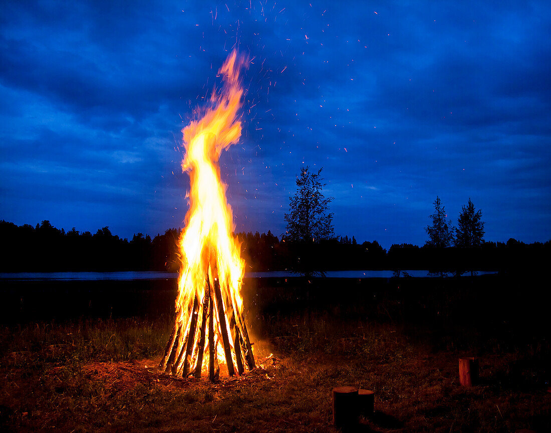 A tall bonfire with flames leaping up, at night. Silhouettes of trees and landscape.
