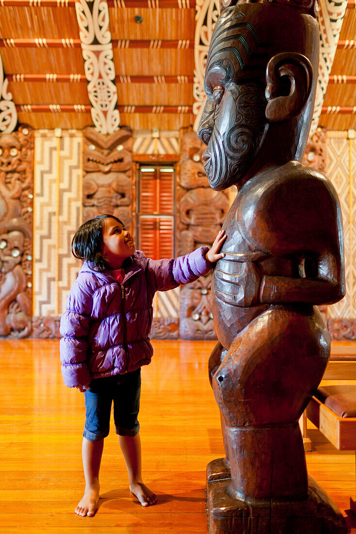 Kleines Mädchen berührt geschnitzte Holzfigur der Ahnen, Stütze im Versammlungshaus in Waitangi Museum, Tatoo Spiralen, Maori Kultur, Nordinsel, Neuseeland