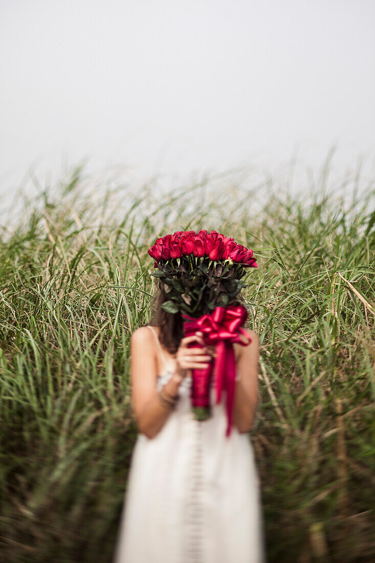 Young Woman Holding Bouquet of Red Roses in Front of Face in Grassy Field