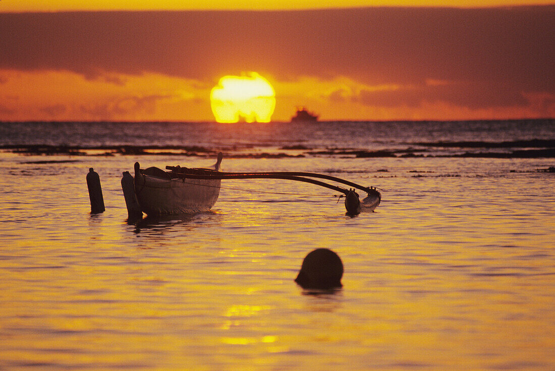 Hawaii, Outigger canoe silhouetted on ocean at sunset, orange reflections on water and sun on horizon.
