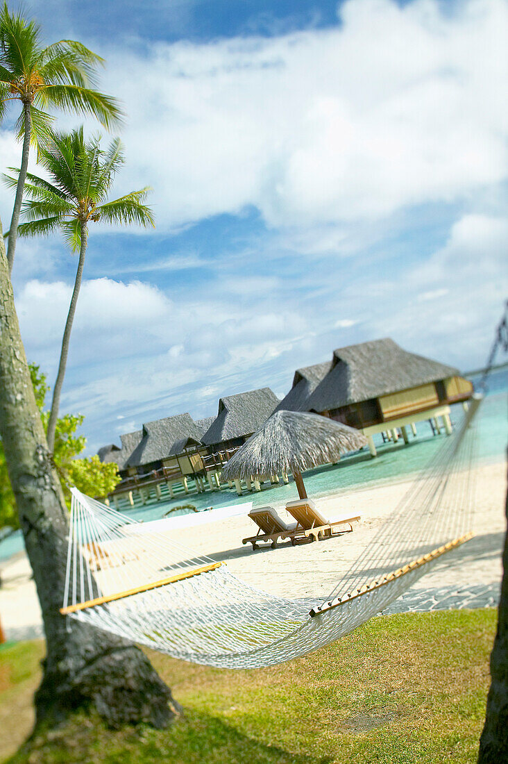 French Polynesia, Tahiti, Bora Bora, hammock in foreground of lounge chairs and thatch umbrella on beach with tranquil ocean and bungalows