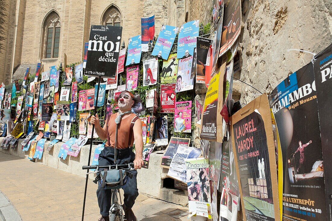 Vaucluse - Festival of Avignon 2012 - Street of the vice légat - The clown Vulcano passes in bike in front of a wall of posters of the shows of the festival Off - In the background: the palace of the Popes