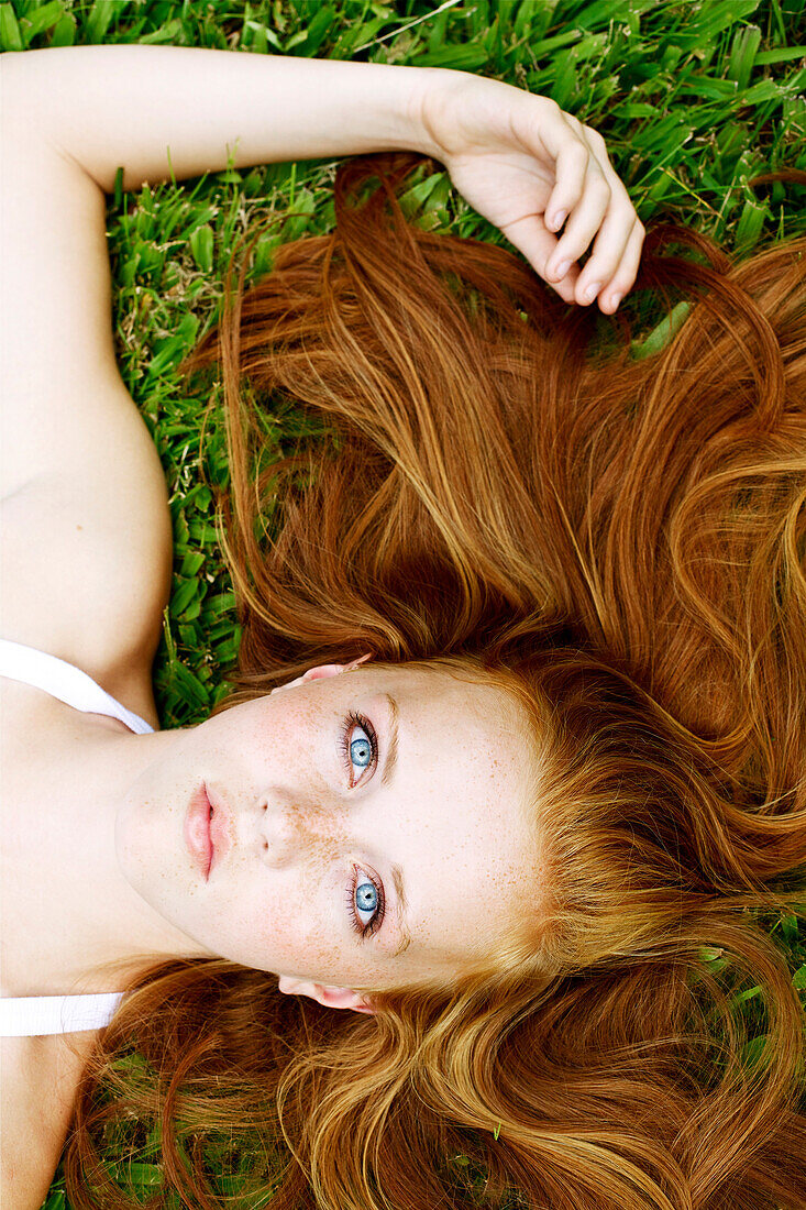 Young woman with freckles and red hair lays in the grass and looks up.