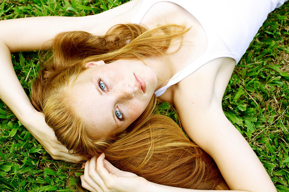 Young woman with freckles and red hair lays in the grass and looks up.