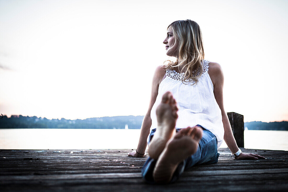 Junge Frau sitzt auf einem Steg in der Abenddämmerung am Starnberger See, Bayern, Deutschland