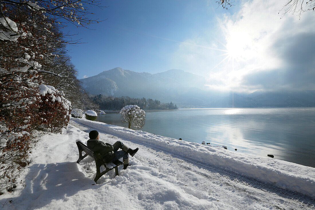 Mann sitzt auf einer Bank an verschneiter Uferpromenade am Kochelsee, Kochel, Oberbayern Deutschland