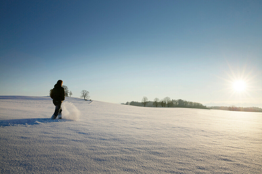 Mann wandernt durch Schnee zur Pfarrkirche St. Johann Baptist, Holzhausen, Münsing, Bayern, Deutschland