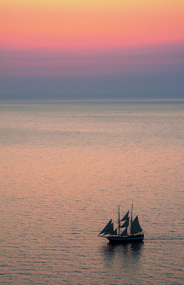 Big masked sailboat sailing in sunset of beautiful Oia in Santorini Greece in Greek Islands from above on cliff into water