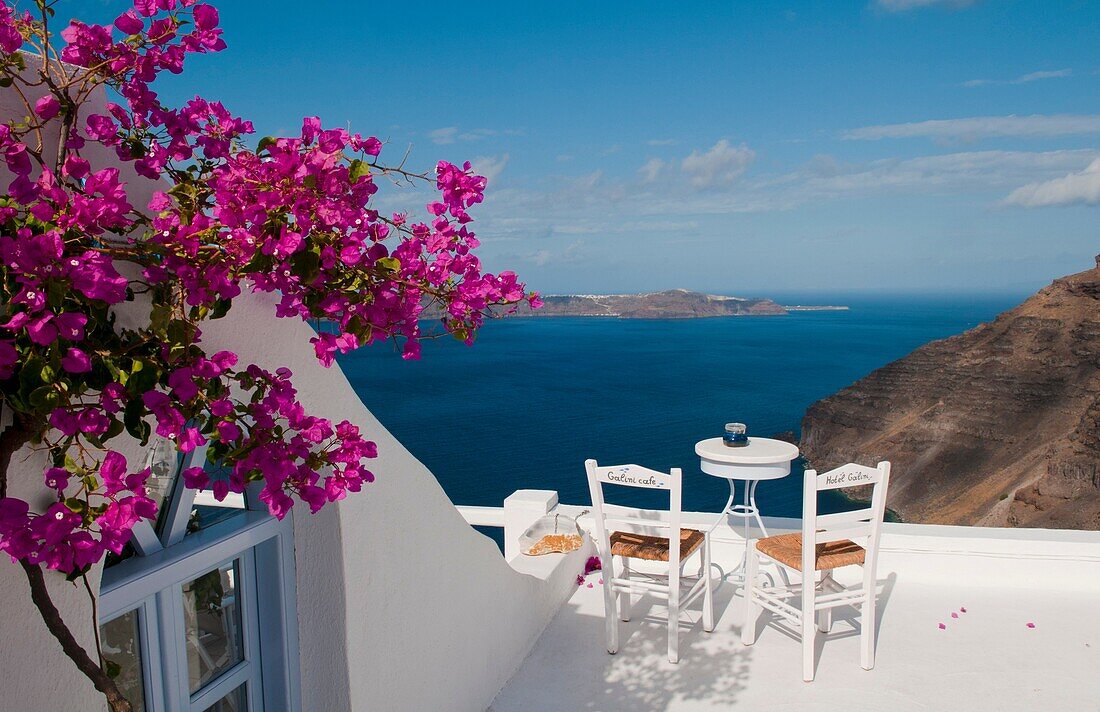 Postcard scene of two lonely chairs and pink flowers on terrace with table ready for tourists in Santorini Greece in Greek Islands
