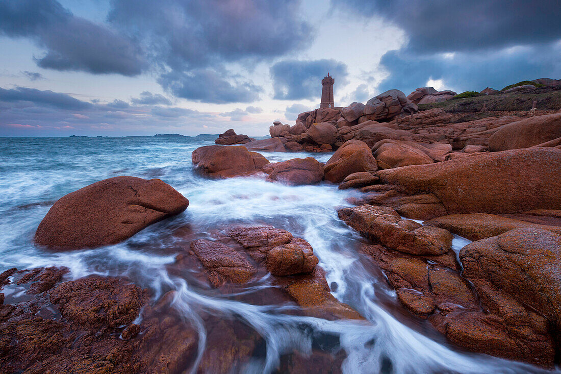 Mean Ruz lighthouse in blue light at dusk with typical red stones of the Cote de Granit Rose in the foreground, Brittany, France