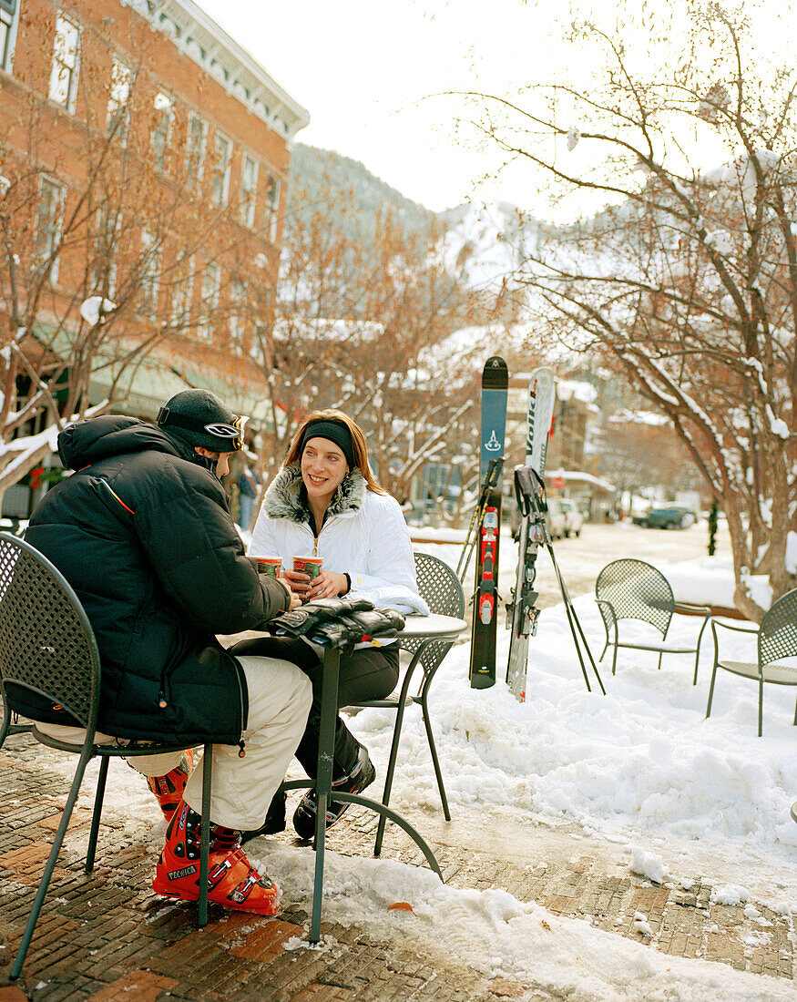 USA, Colorado, Aspen, couple wearing ski gear having coffee at an outdoor table in the winter, Downtown