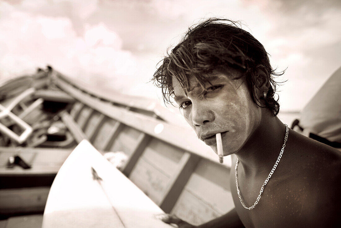 INDONESIA, Mentawai Islands, Kandui Surf Resort, young surfer with sunscreen on his face, smoking and holding a surfboard (B&W)