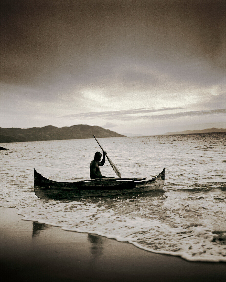 MADAGASCAR, Nosy Komba, Jardin Vanille, man canoeing in a dugout canoe at dusk (B&W)