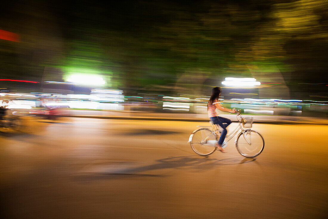 VIETNAM, Hanoi, a young woman rides her bike down a busy street at night, Hoan Kiem Lake in the distance