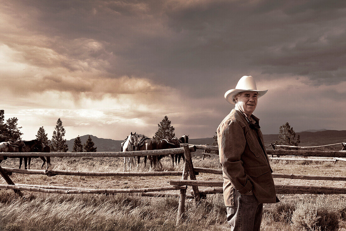 USA, Wyoming, Encampment, a man in a cowboy hat stands next to a fenceline with horses and an ominous sky in the distance