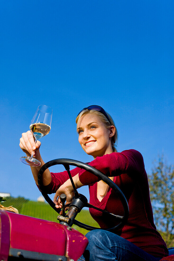 Junge Frau sitzt mit einem Glas Weißwein auf einem Traktor, Steiermark, Österreich