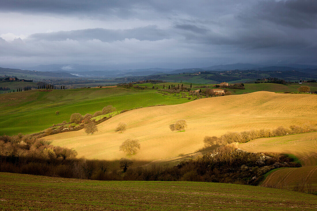 Toskanische Hügellandschaft des Val d'Orcia im Frühling mit Sonne im Vordergrund und Regenwolken im Hintergrund, San Quirico d’Orcia, Toskana, Italien