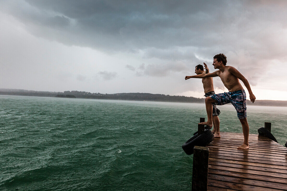 Two young men on a jetty leaning into the wind, Lake Starnberg, Bavaria, Germany