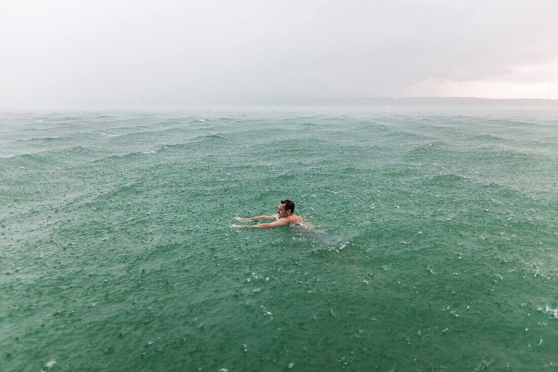 Junger Mann schwimmt bei Regen im Starnberger See, Bayern, Deutschland