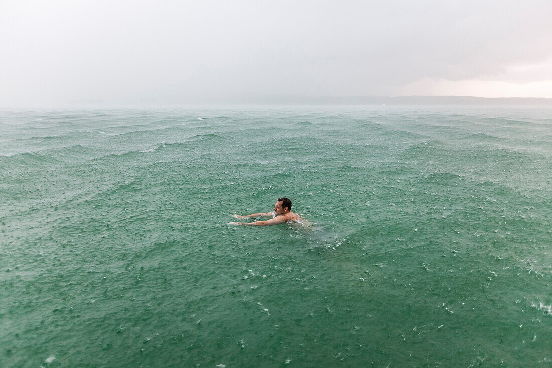 Young man swimming in Lake Starnberg in rain, Bavaria, Germany