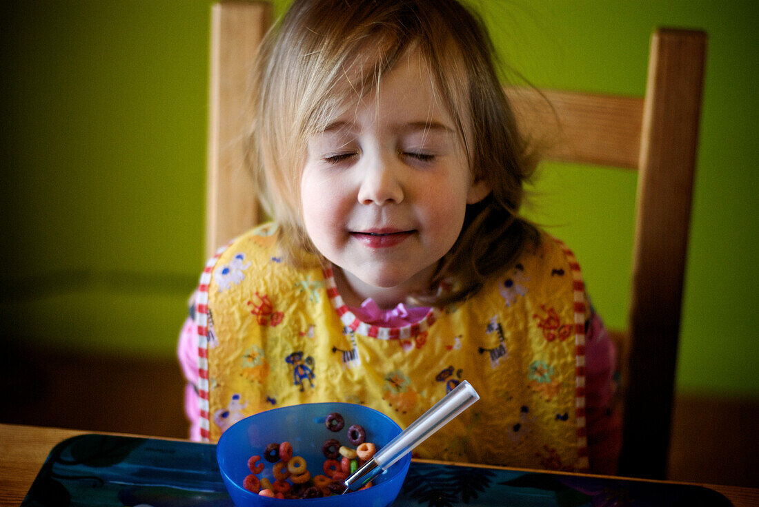 Little girl at table with bowl of cereal and eyes closed, Otterburn, Quebec