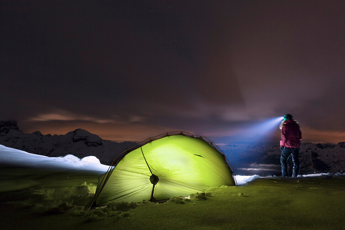 Young woman with headlamp standing beside a tent in snow, the Dolomites, Belluno, Veneto, Italy