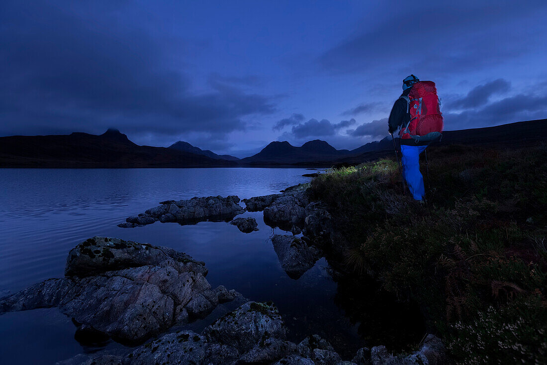 Junger Mann wandert am Loch Bad a Ghaill in der Dämmerung, Stac Pollaidh, Cul Beag, Sgorr Tuath und Ben Mor Coigach im Hintergrund, Assynt, Schottland, Großbritannien