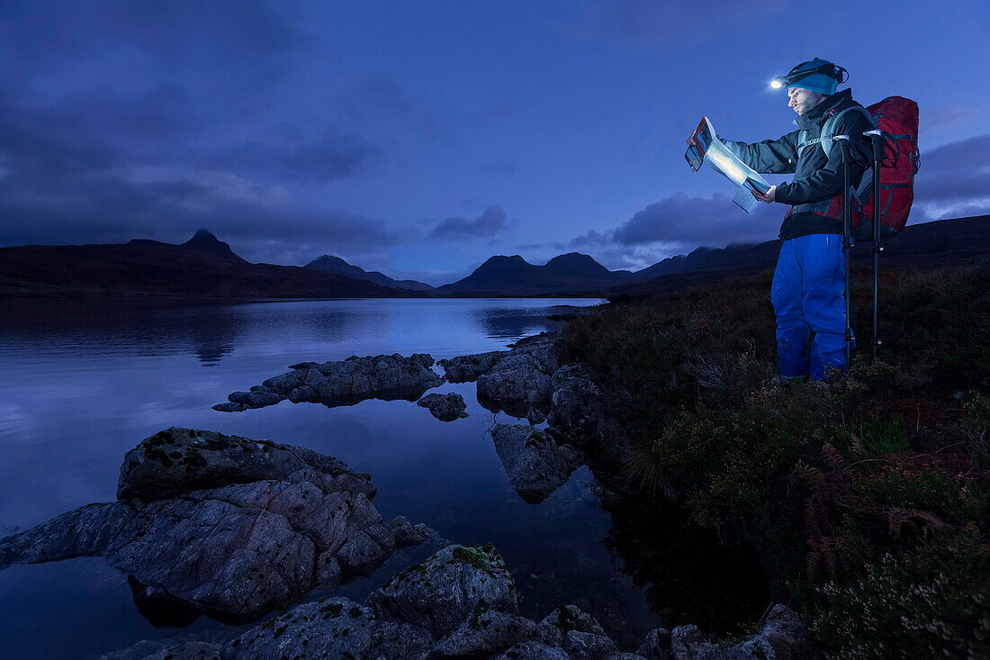 Junger Mann mit Stirnlampe liest eine Wanderkarte am Loch Bad a Ghaill in der Dämmerung, Stac Pollaidh, Cul Beag, Sgorr Tuath und Ben Mor Coigach im Hintergrund, Assynt, Schottland, Großbritannien