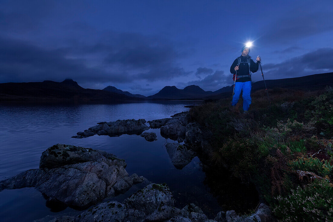 Junger Mann mit Stirnlampe wandert am Loch Bad a Ghaill in der Dämmerung, Stac Pollaidh, Cul Beag, Sgorr Tuath und Ben Mor Coigach im Hintergrund, Assynt, Schottland, Großbritannien