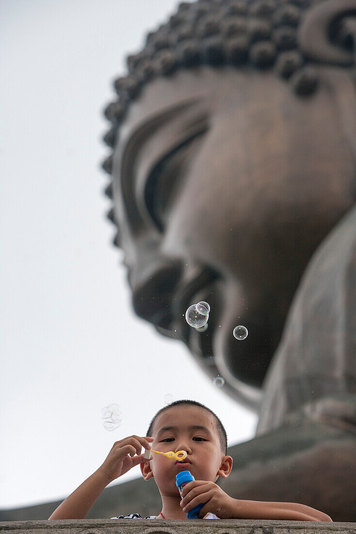 Kleiner Junge pustet Seifenblasen unterhalb der riesigen Statue des Giant Buddha auf dem Ngong Ping Plateau der Insel Lantau, Hong Kong, China, Asien