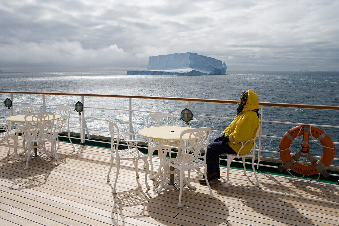 Frau in Winterkleidung enstpannt sich an Deck von Kreuzfahrtschiff MS Deutschland (Reederei Peter Deilmann) mit Eisberg im Hintergrund, nahe King George Island, Südliche Shetland-Inseln, Antarktis