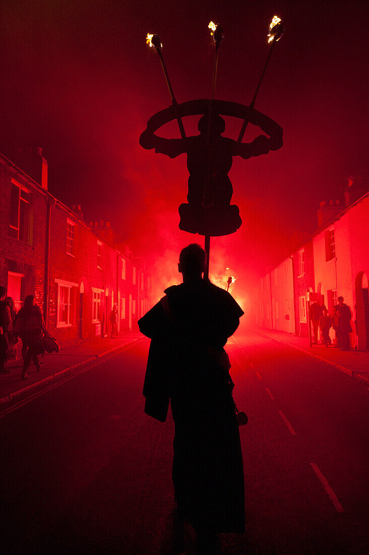 Silhouettes Of Monk With Banner Lit By Red Flare Walking Through Smokey Backstreets Of Lewes On Bonfire Night, East Sussex, Uk
