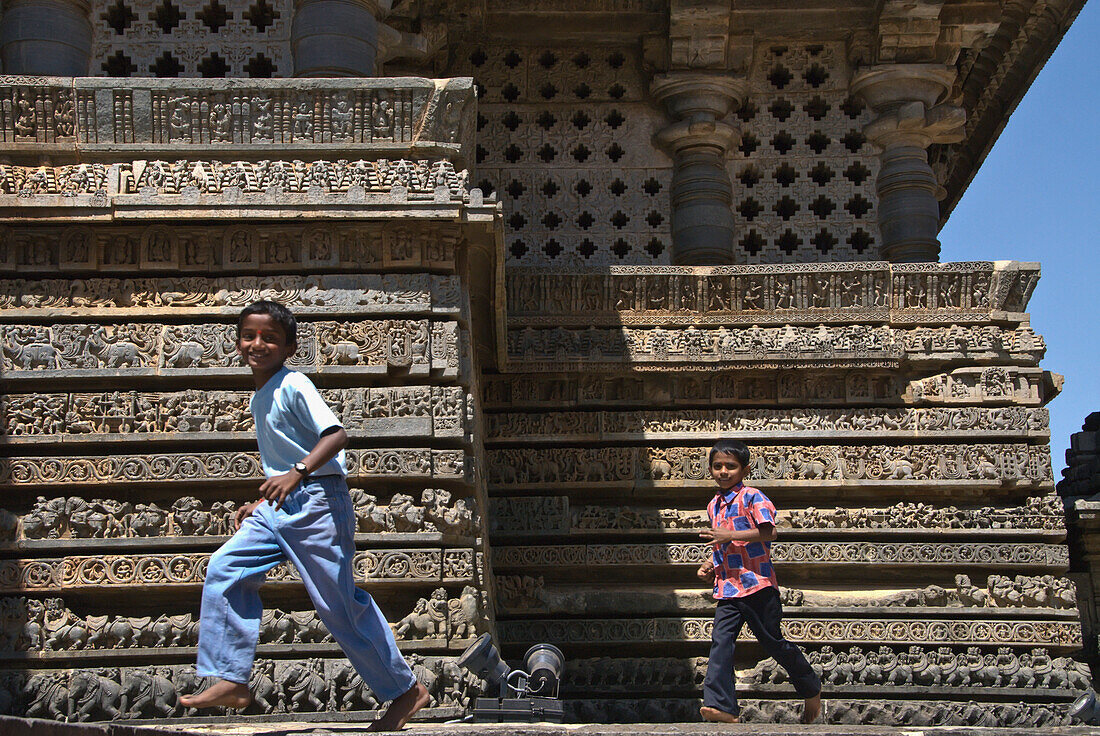 Two Boys Running In Front Of Ornate Carvings On Halebid Temple, Halebid, Karnataka, India