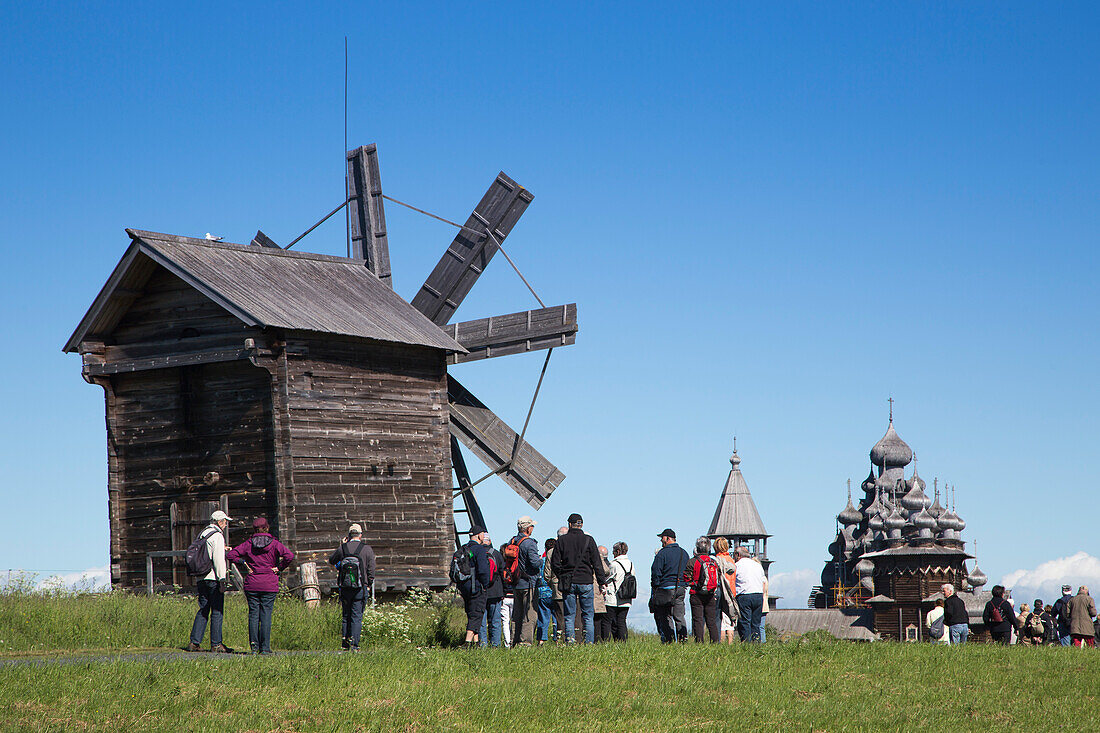 People in front of wooden windmill, belfry and Church of the Intercession of the Virgin and Church of the Transfiguration at Kizhi Pogost in distance, Kizhi Island, Lake Onega, Russia, Europe