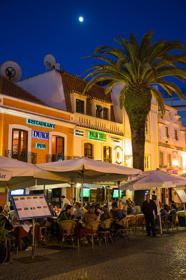 Menschen sitzen draußen vor Restaurants an der Praca Costa Pinto in der Abenddämmerung, Cascais, nahe Lissabon, Portugal