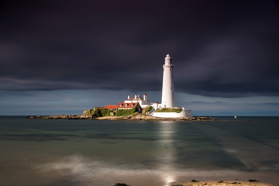 'White Lighthouse Illuminated By Sunlight Under A Dark Stormy Sky; St. Mary's Island Northumberland England'