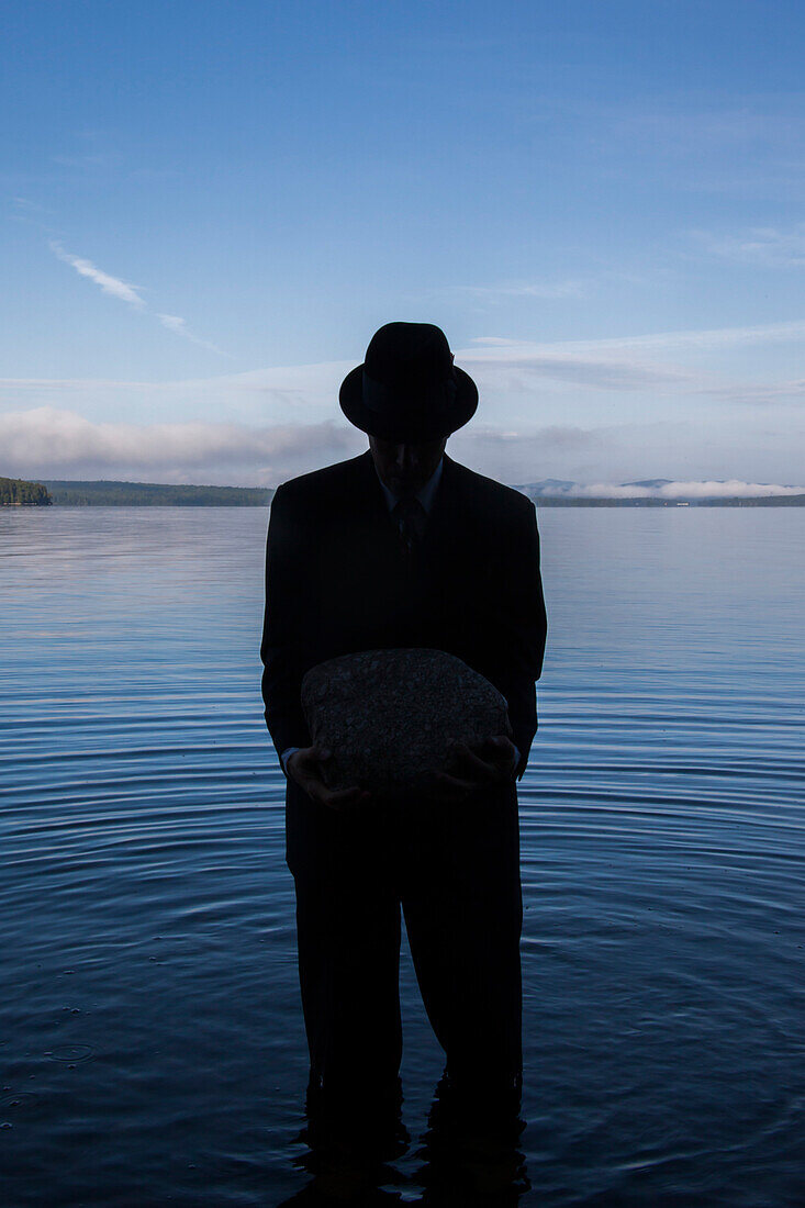 Businessman in Suit and Hat Holding Boulder in Lake