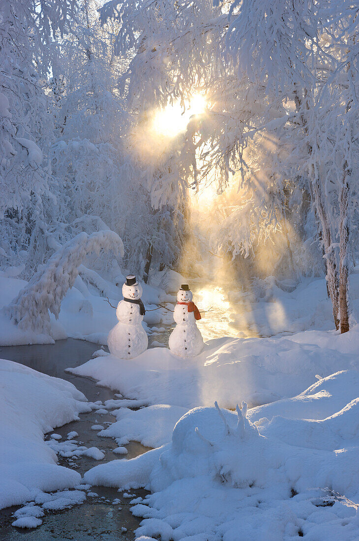 Snowman couple standing next to a stream with sunrays shining through fog and hoar frosted trees in the background, Russian Jack Springs Park, Anchorage, Southcentral Alaska, Winter. Digitally enhanced.
