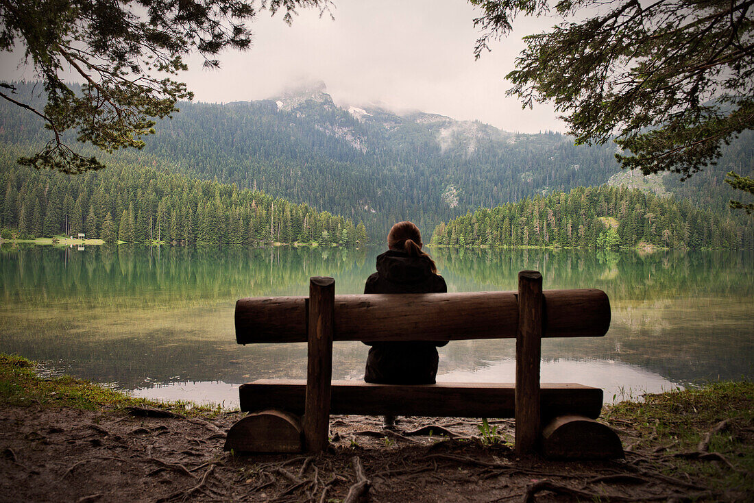 Young woman sitting on a bench looking across the Black Lake (Crno jezero) in Durmitor National Park, Zabljak, Montenegro, Western Balkan, Europe