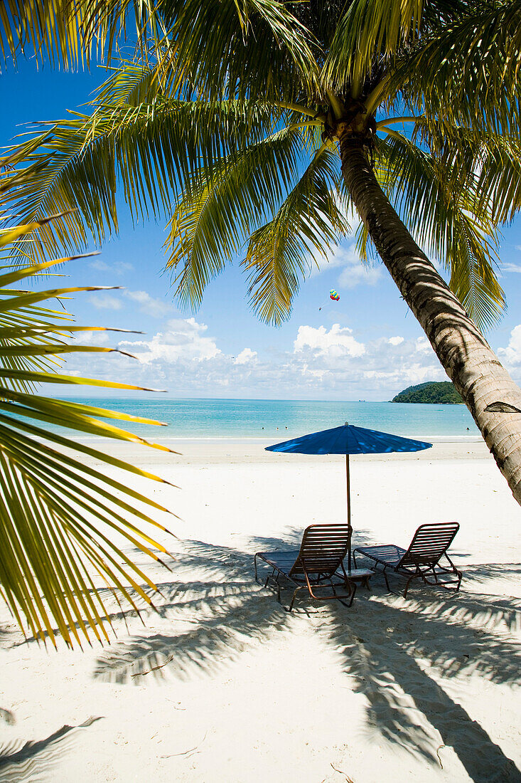 Deck chairs under parasol on white sandy beach with palm trees overlooking blue sea, Pantai Cenang (Cenang beach), Pulau Langkawi, Malaysia