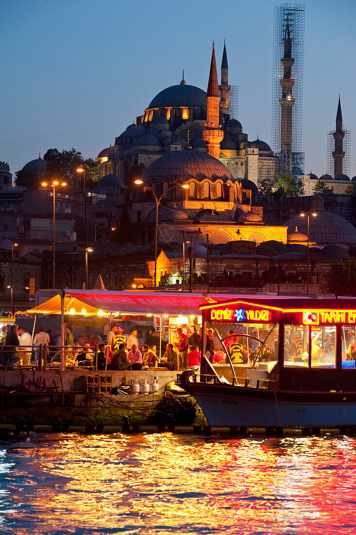 Fish restaurants being run from side of boats beside Golden Horn with Suleymaniye Mosque behind at dusk, Istanbul, Turkey