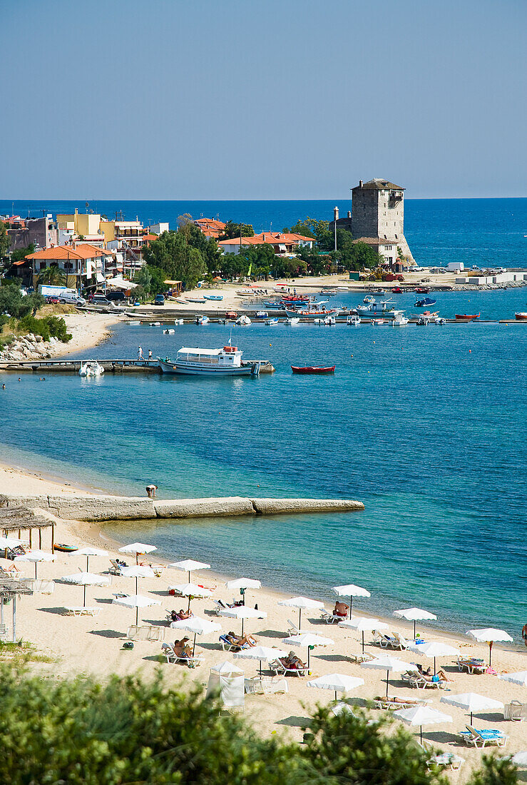 Tourist beach with beach umbrellas and sun bathers with Byzantine tower, Tower of Prosforios in the distance, Ouranoupoli, Halkidiki, Greece