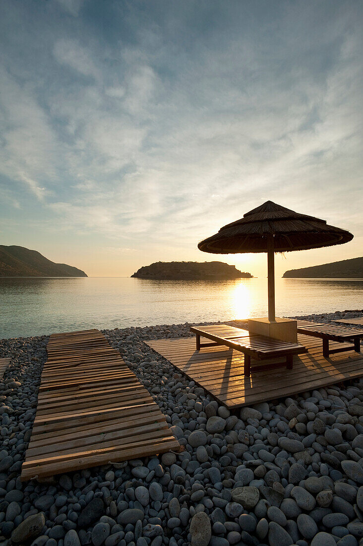 Umbrella and sun loungers on beach opposite Spinalonga Island at dawn, Crete, Greece